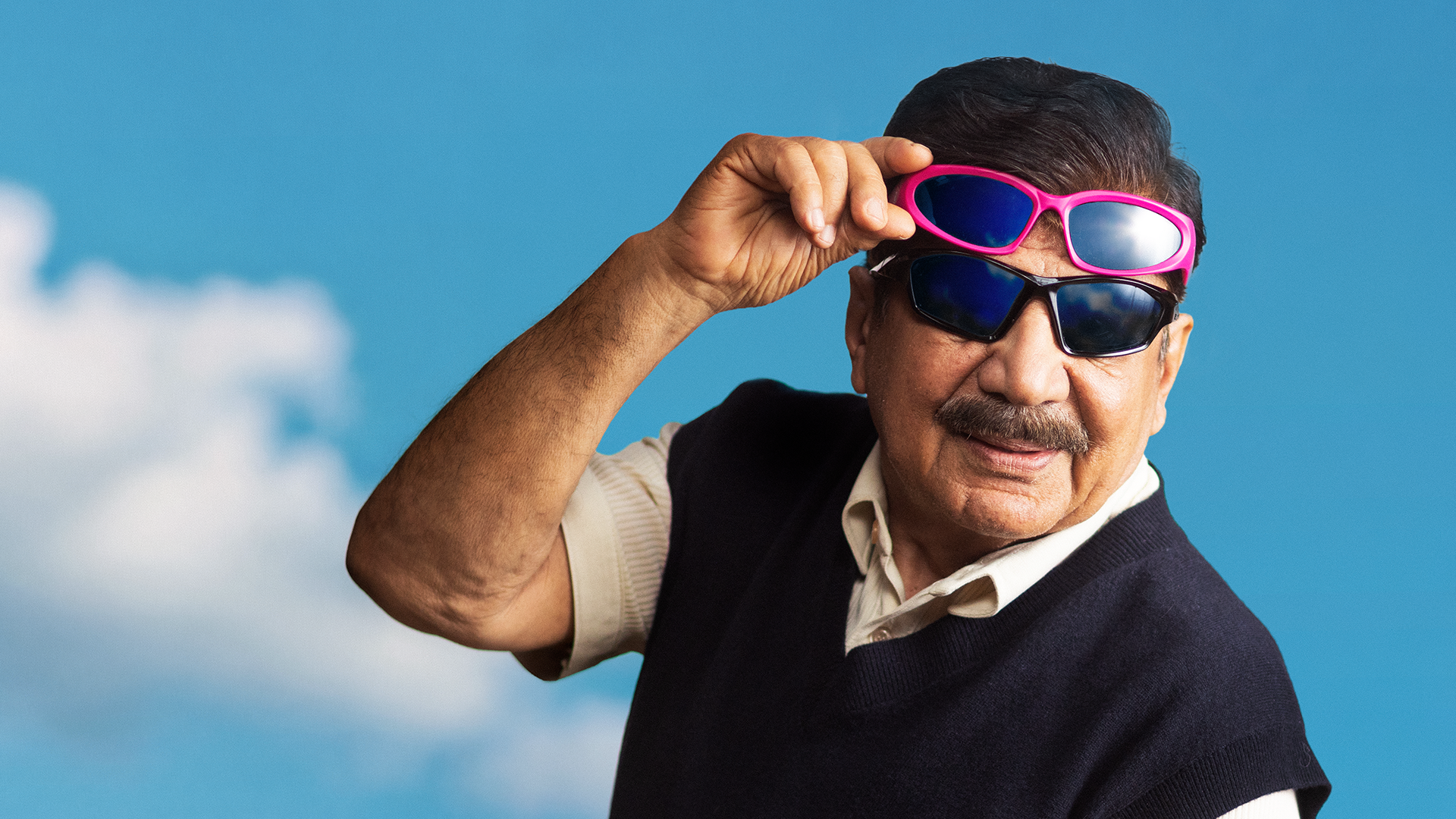 En äldre man med mustasch bär två par solglasögon utomhus mot en blå himmel med moln.