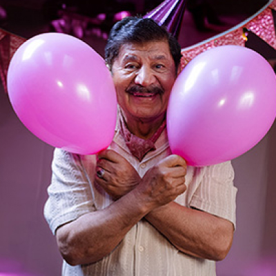 En leende äldre man med mustasch håller två rosa ballonger och bär en festmössa vid en fest.