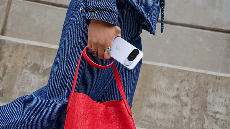 En hand med ringar håller en vit Google Pixel 10 och en röd läderväska, klädd i blå denimkläder.