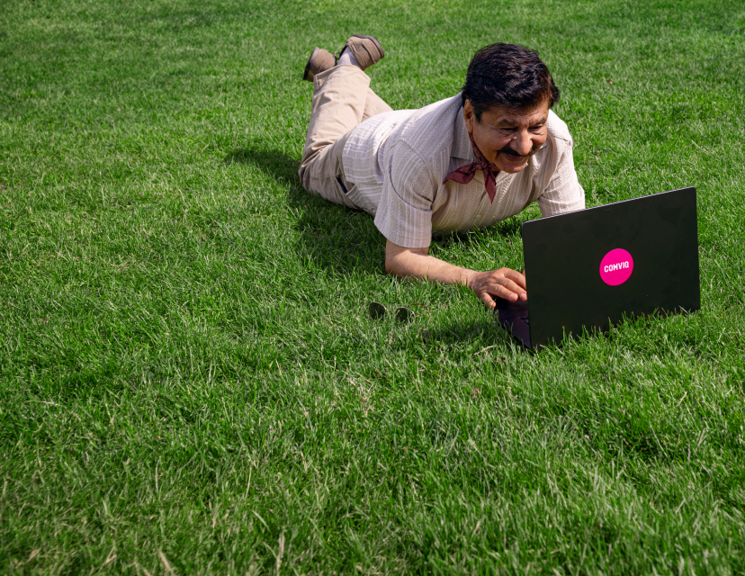 En leende äldre man med mörkt hår och mustasch ligger på grönt gräs och använder en svart laptop med en rosa Comviq-klistermärke.