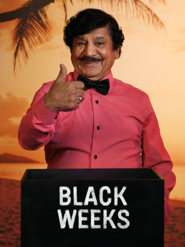 Person i rosa skjorta gör tummen upp framför tropisk solnedgång med palmer, över en "BLACK WEEKS"-skylt.
