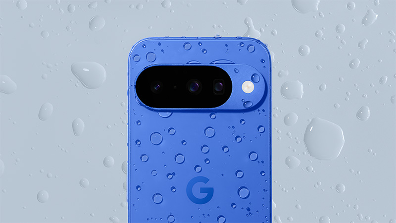 Blå Google Pixel 10 med trippelkamera täckt av vattendroppar mot en ljusgrå bakgrund.