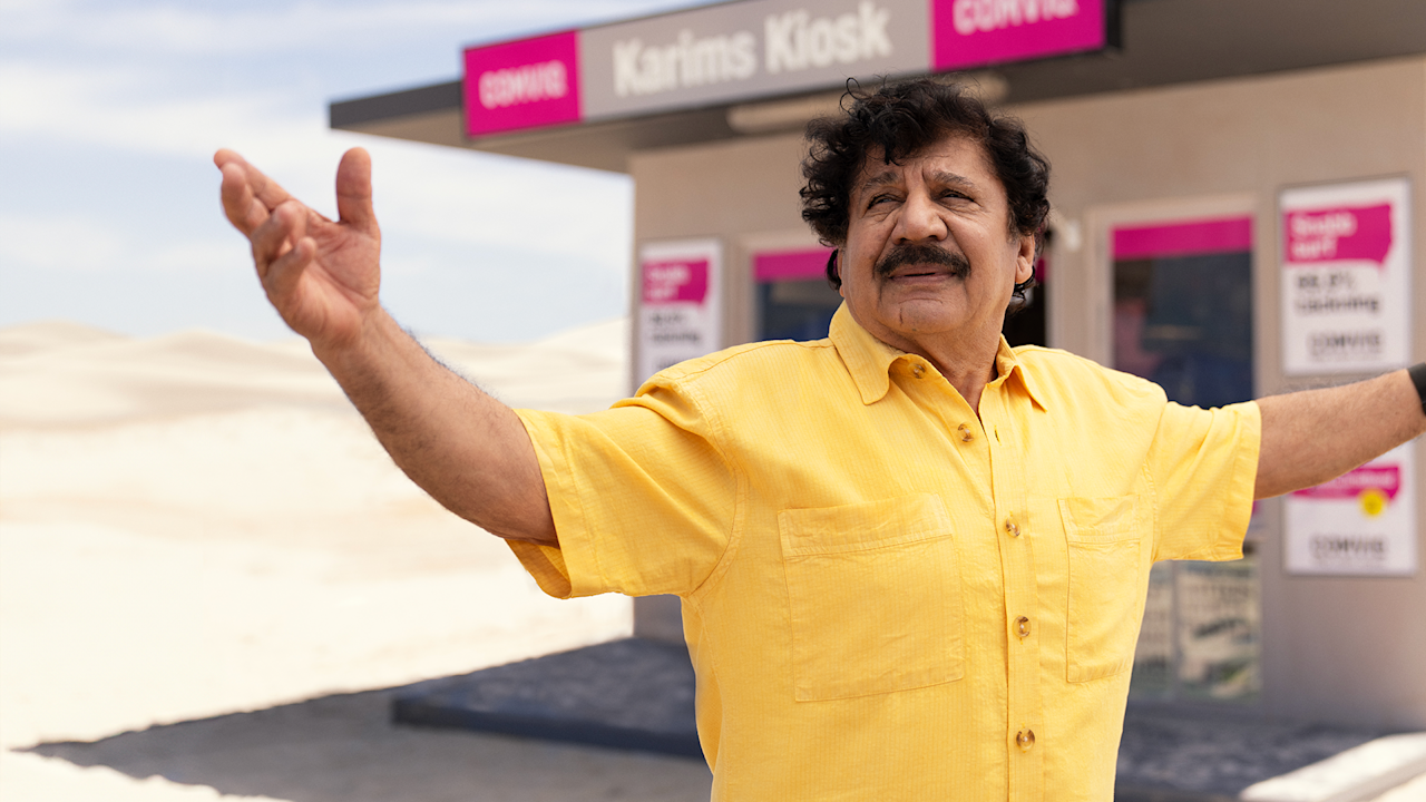 Karim utanför kiosk, armar utsträckta i omfamnande rörelse, gul skjorta, ökenmiljö, i bakgrunden skylt med Comviq-logotyp och texten "Karims Kiosk"