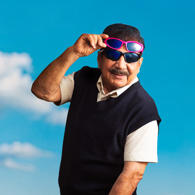 En äldre man med mustasch bär två par solglasögon utomhus mot en blå himmel med moln