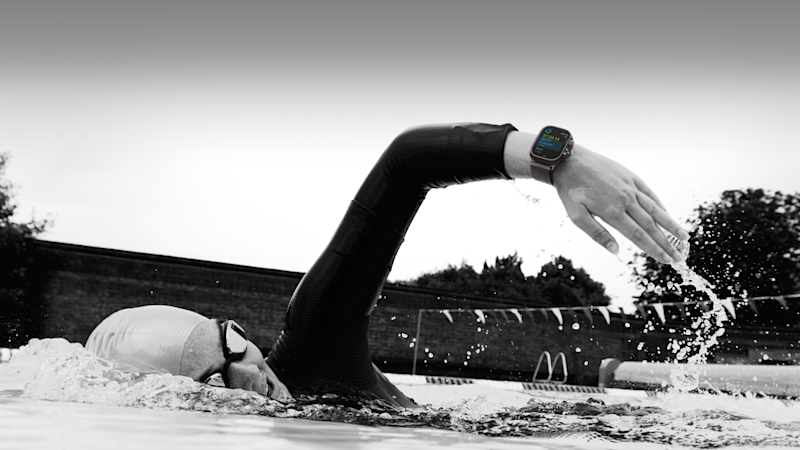 Simmare i våtdräkt med Apple Watch Ultra 3 tränar crawl i utomhuspool, svartvit bild med vattenstänk.