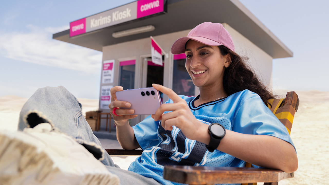 Nadja sitter i stol och tittar på sin mobil utanför Comviq-kiosken, rosa mobil och smartwatch på handleden, i bakgrunden ökenmiljö