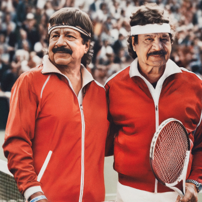 Två äldre män, båda har mörkt hår och mustasch, i röda träningsjackor och pannband på en tennisbana. En håller ett racket. Publik i bakgrunden.