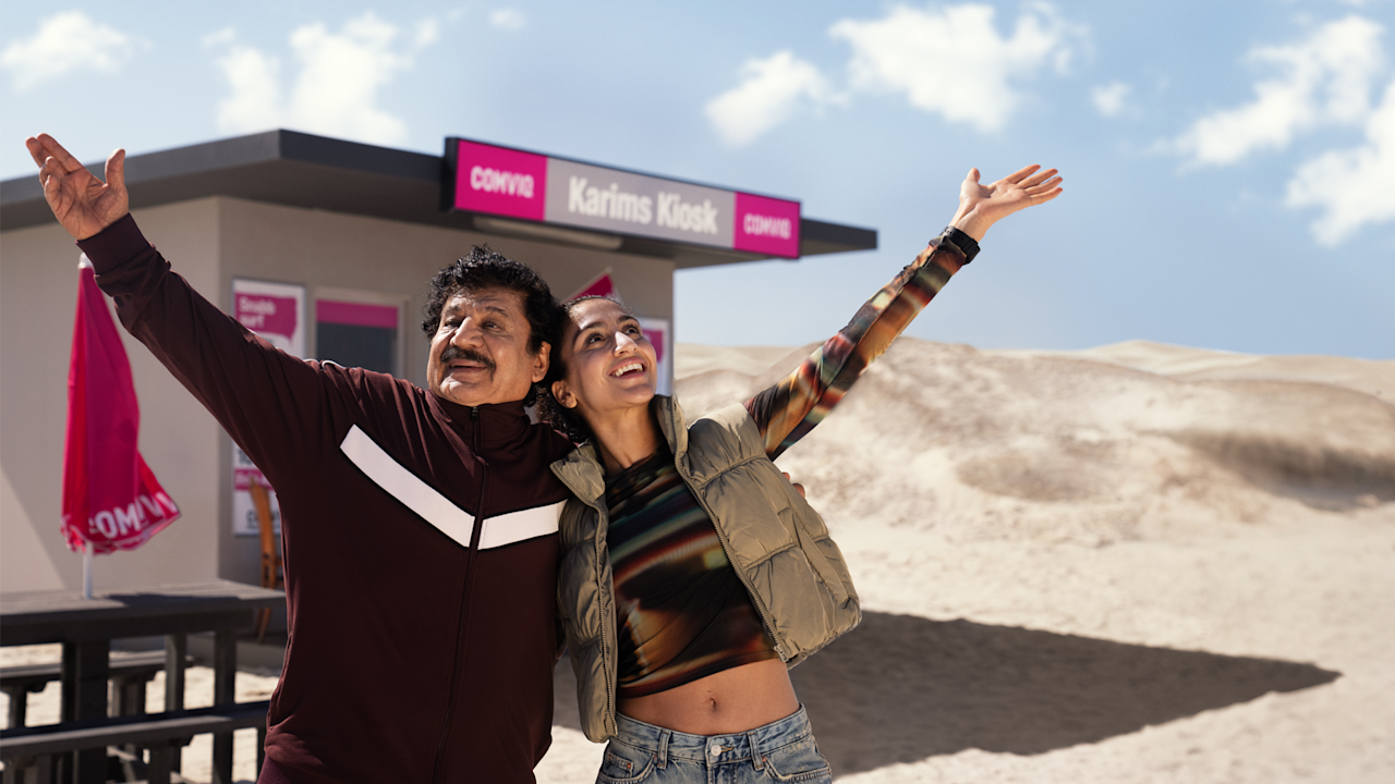 Karim och Nadja sträcker upp händerna mot himlen och ler, kiosk i bakgrunden, skylt med texten "Karims Kiosk" och Comviq-logotyp