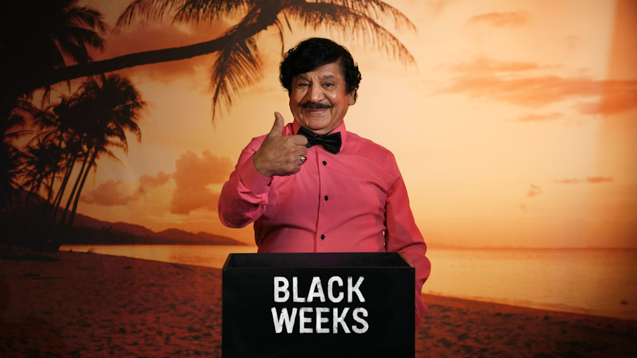 Person i rosa skjorta och fluga visar tummen upp framför en låda med texten "BLACK WEEKS" mot solnedgångsbakgrund med palmer.