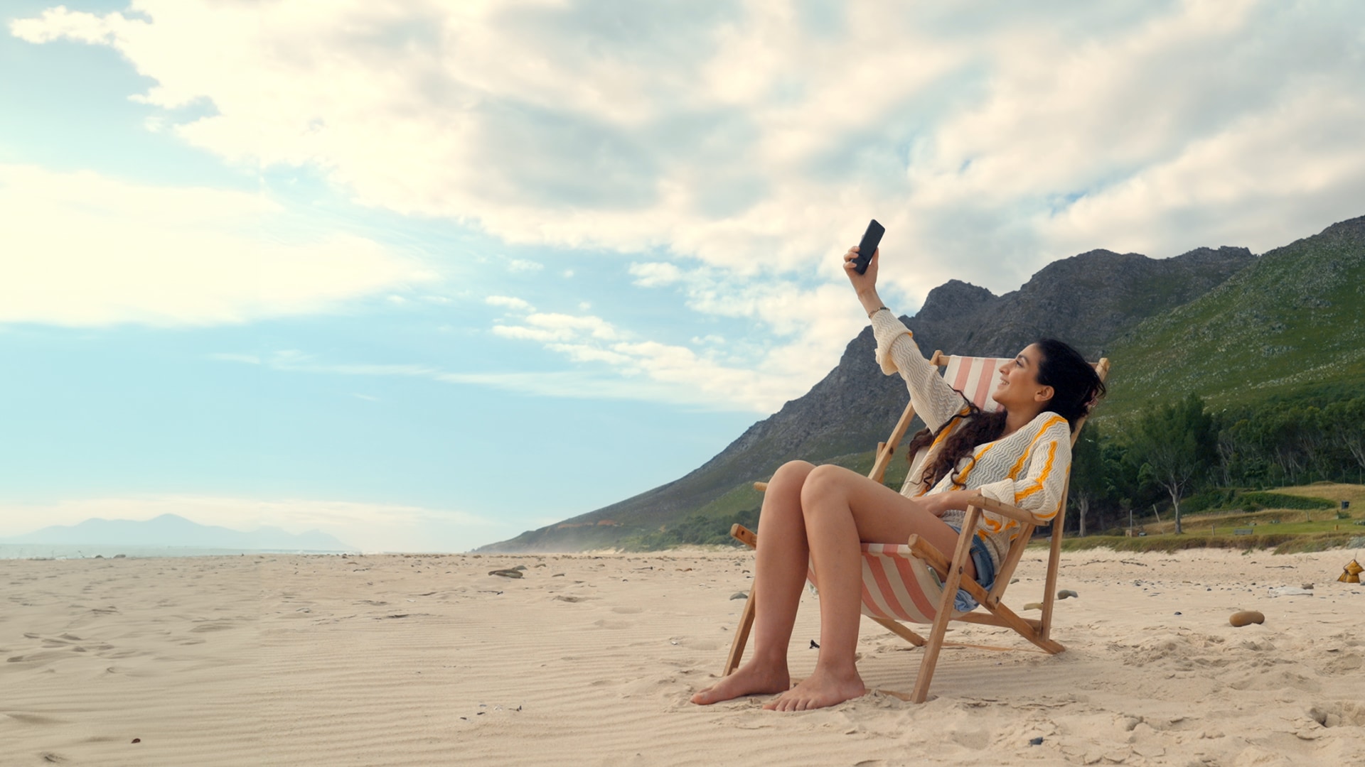 Nadja sitter i solstol på stranden och tar en selfie, lång sandstrand, grönklädda berg, blå himmel och vita moln