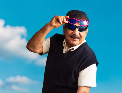 En äldre man med mustasch bär två par solglasögon utomhus mot en blå himmel med moln