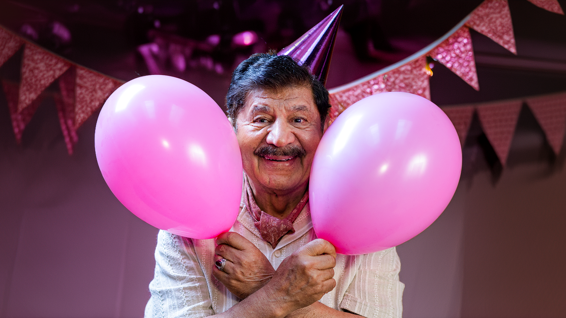 En leende äldre man med partyhatt håller två rosa ballonger omgivna av rosa vimplar i bakgrunden.