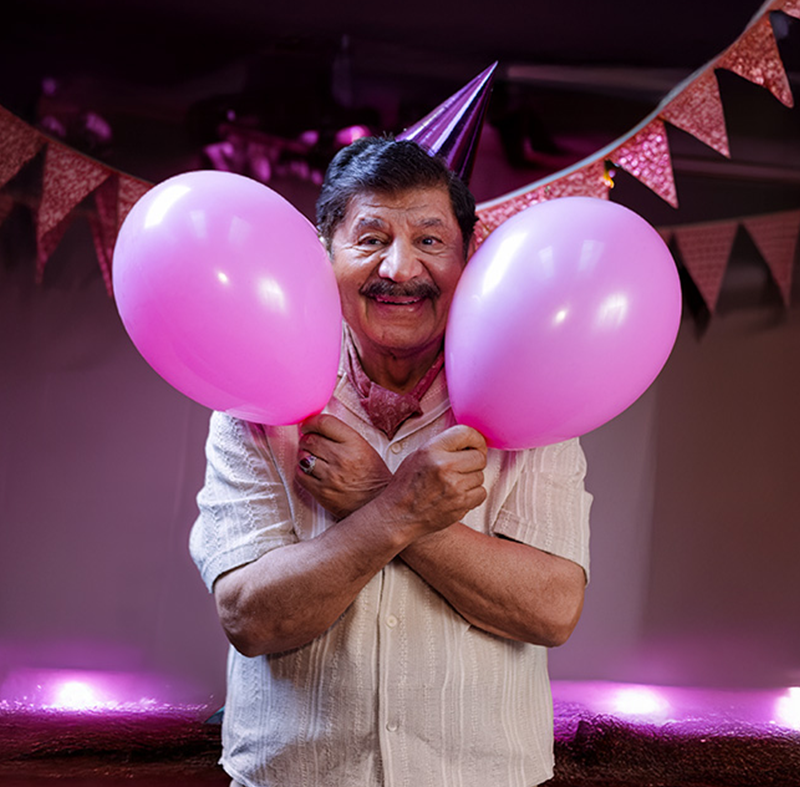 En leende äldre man med partyhatt håller två rosa ballonger omgivna av rosa vimplar i bakgrunden