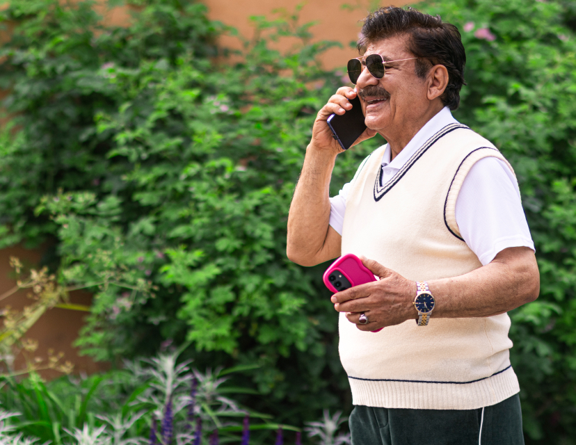 En äldre mörkhårig man med mustach och solglasögon pratar i mobiltelefon utomhus, håller en rosa telefon i andra handen, grön bakgrund.