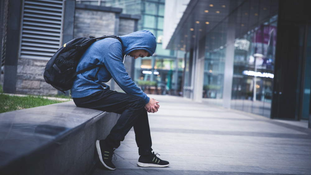 Youth unemployment A young man wearing a blue hoodie and black backpack sits hunched forward on a concrete ledge outside a modern office building