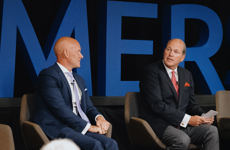 two men sitting and speaking to a crowd