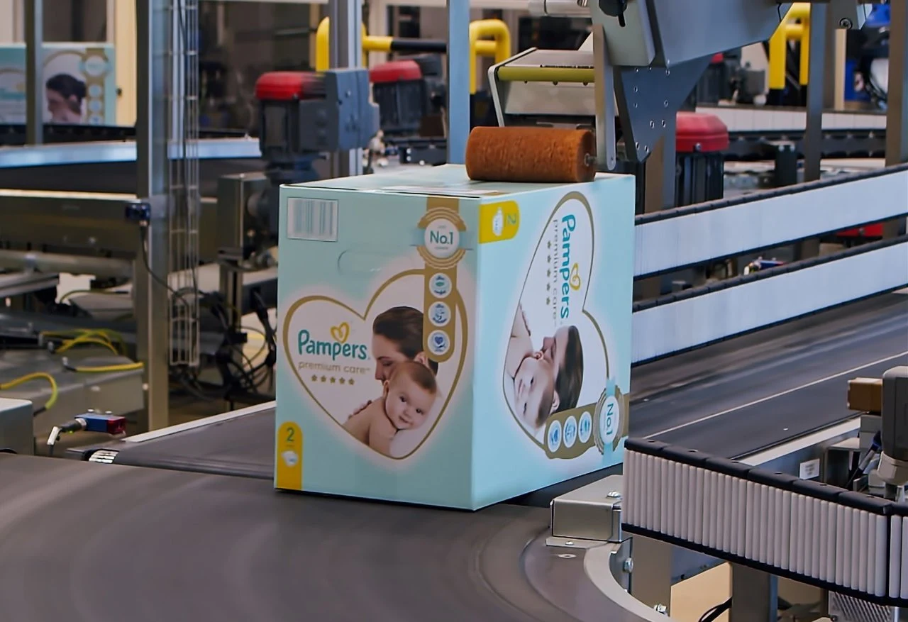 Niebieskie pudełko pieluszek Pampers Premium Care na taśmie produkcyjnej w fabryce z maszynami pakującymi w tle.