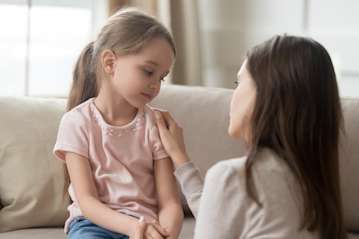 Mother and young daughter sit together on a sofa. The mother has her hand on the girl's shoulder.