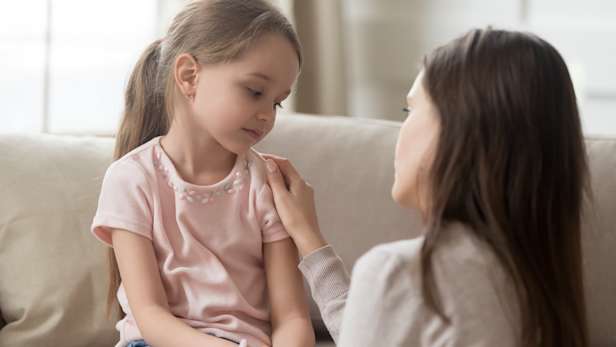 Mother and young daughter sit together on a sofa. The mother has her hand on the girl's shoulder.