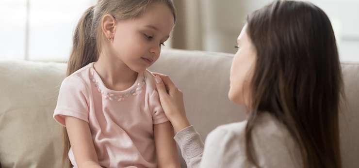 Mother and young daughter sit together on a sofa. The mother has her hand on the girl's shoulder.