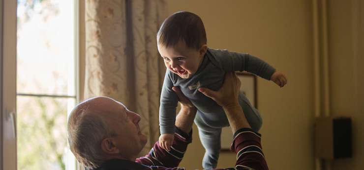 Older person holds a baby in the air by a window