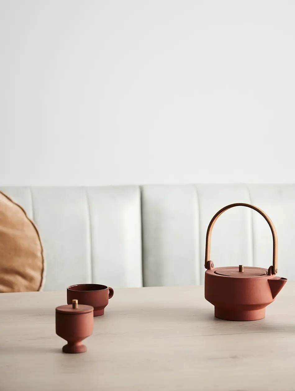 Dining table with bench seat and modern teapot, cup and sugar bowl in neutral colours.