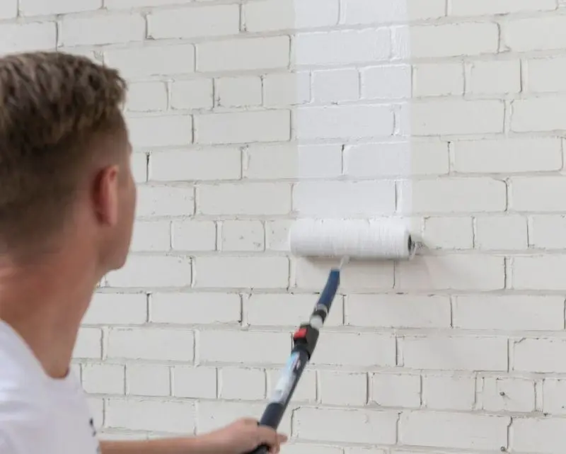 Man rolling a roller with white paint on it on a brick wall