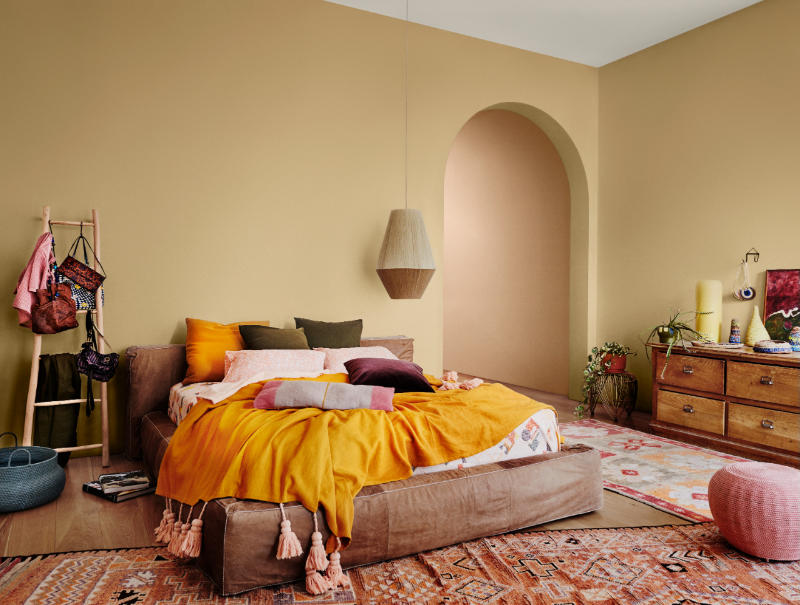 A bedroom with a double bed on the floor and a hanging rattan style lamp to the side of the bed. There is an arched doorway and the walls are painted yellow. There are rugs covering the wooden floor.