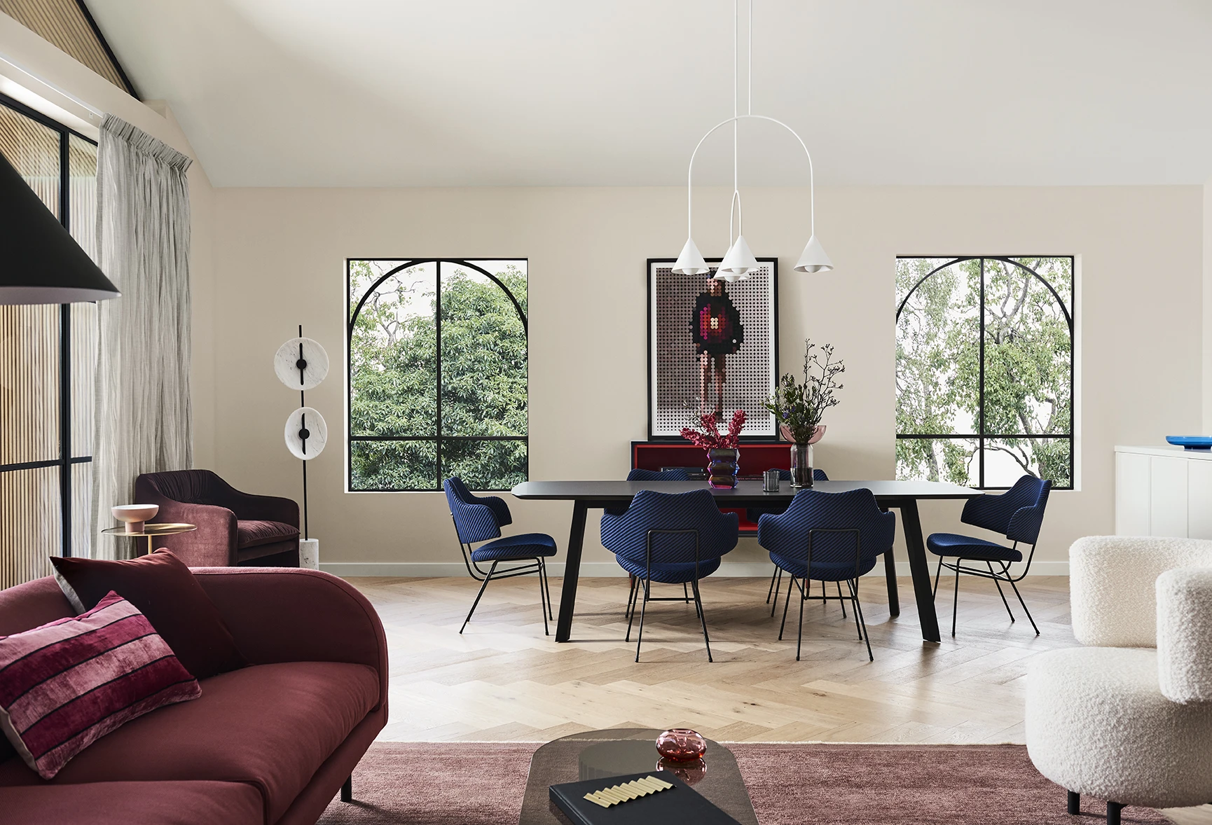 Neutral coloured dining room with black table and blue chairs around it. In the foreground is a red couch in the living room