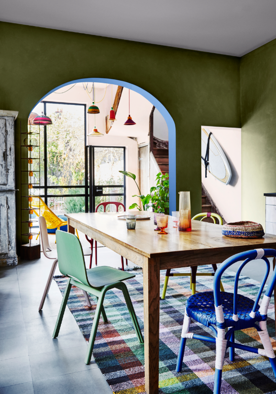 Dining room with coloured cross checkered rug, different style and coloured chairs, a wooden dining table and a green wall with an archway in the background