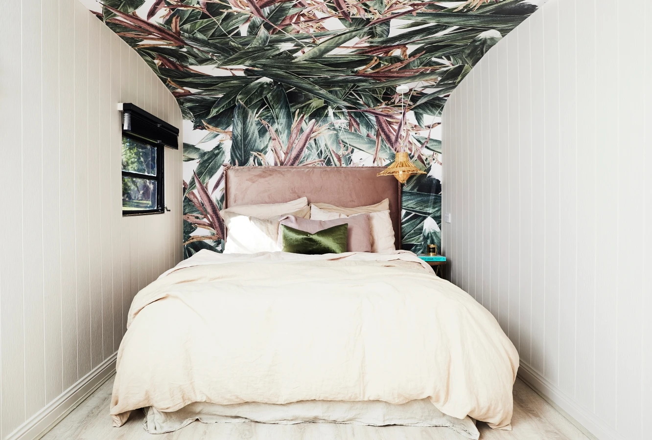 Double bed with pink velvet bedhead in retro caravan. Jungle print wallpaper on ceiling and follows curve of the caravan wall to become feature of the room. Neutral side walls