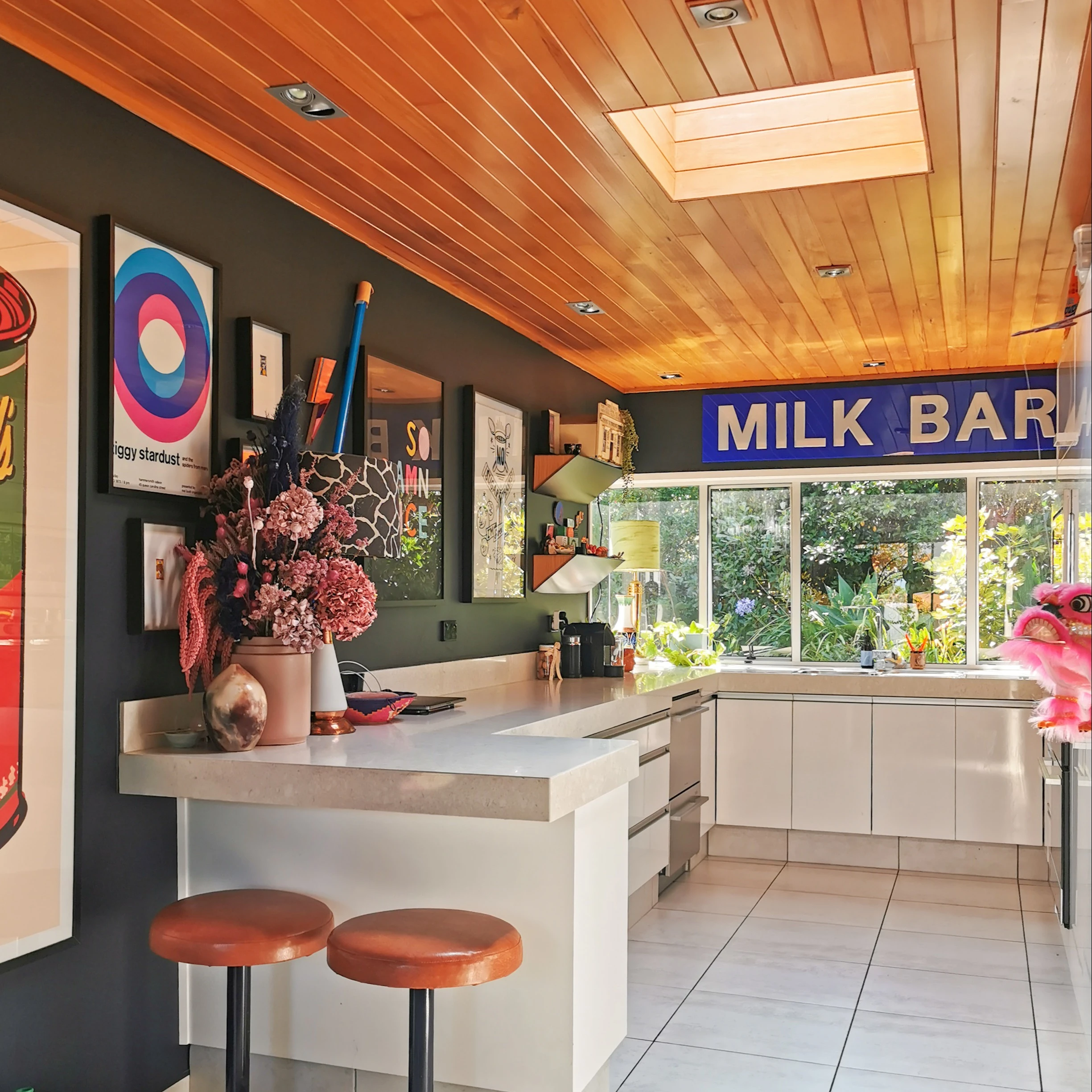 Evie Kemp's kitchen has white tiles on the floor with white cabinets and a white benchtop. Her wall is dark green with lots of colourful artwork on it. The ceiling is wooden and above the bench sits a sign that looks like a street sign that says 'milk bar'