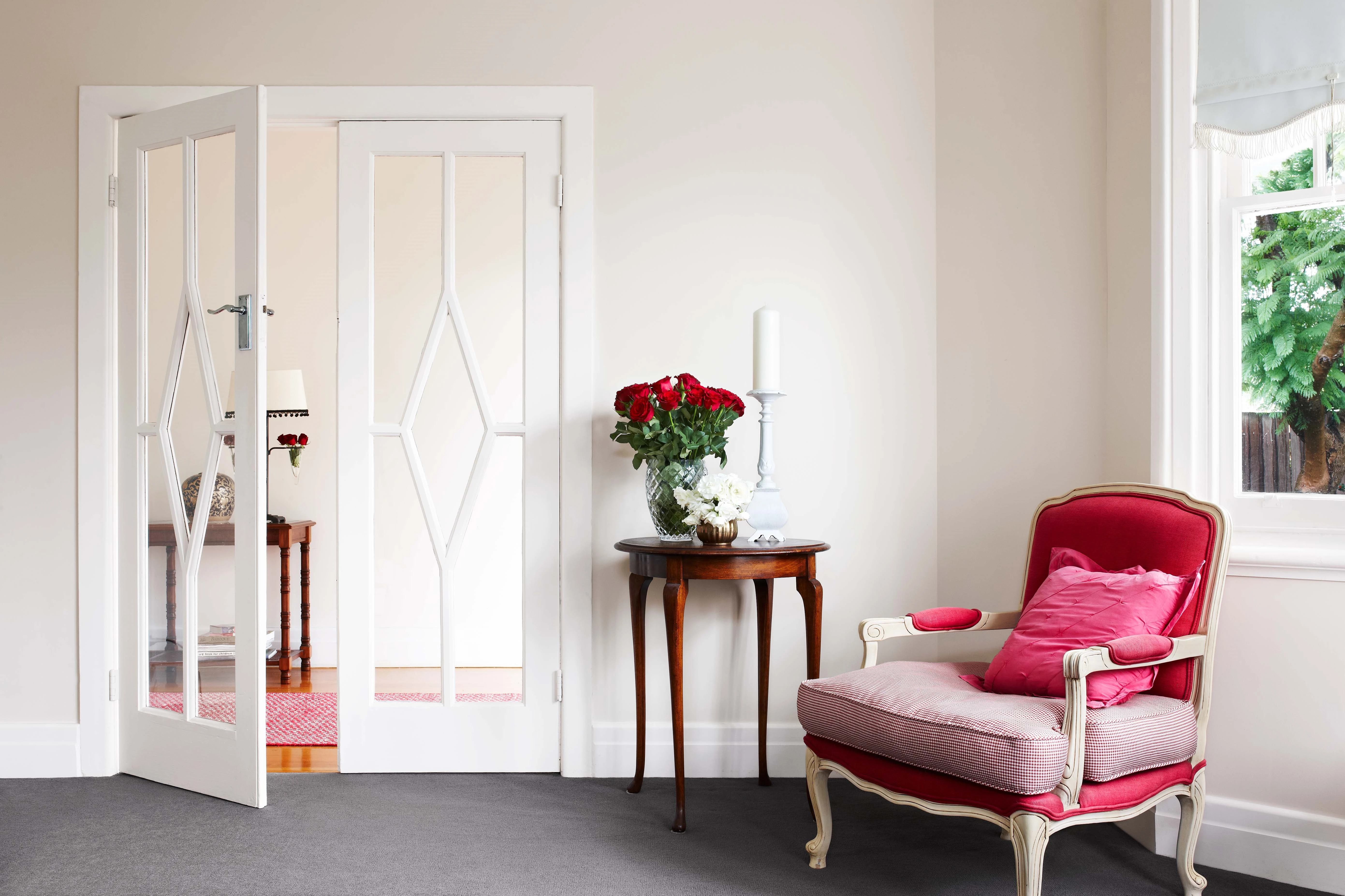 White living room with glass double doors with pink armchair and side table with vase of roses.