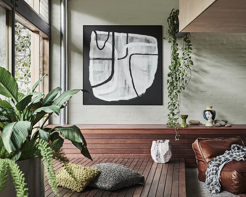 A wooden interior deck with wooden bench seat in front of light green brick wall. There is a large piece of abstract art on the wall and a leather beanbag style seat on the floor.