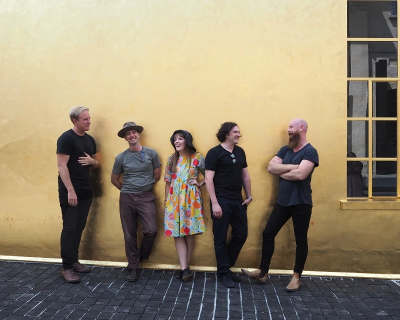 Four men and a woman leaning on a yellow exterior wall.