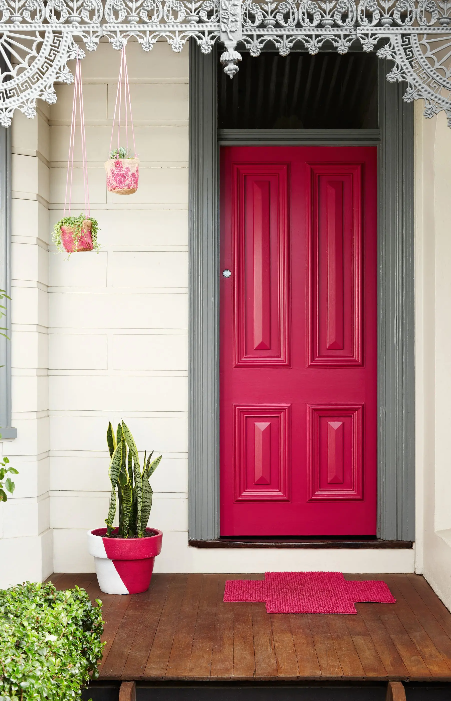Narrow view of bright pink door with matching floor mat. Pot plant sitting beside door and hanging from roof.