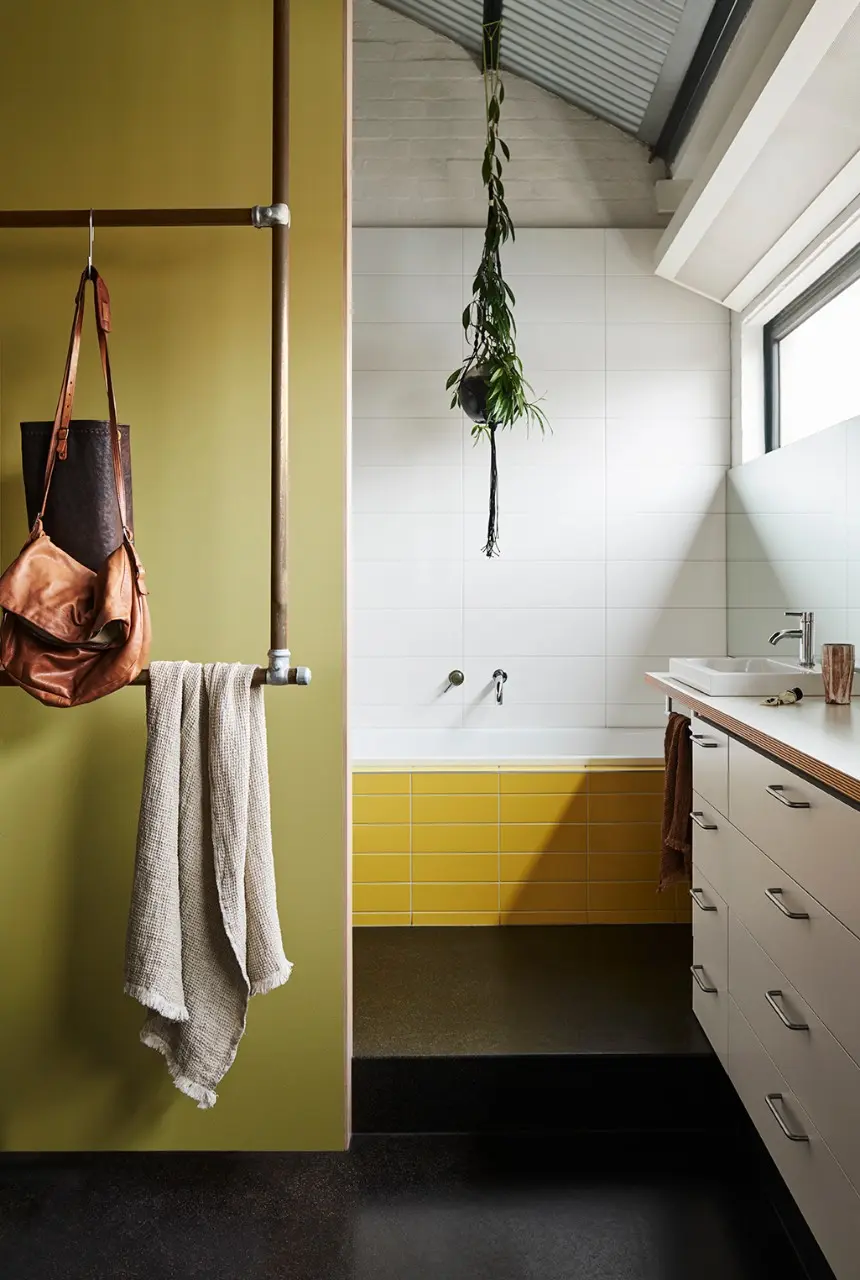 Bathroom with yellow wall and yellow bricked bath. A plant hangs in a pot from the ceiling and there are industrial looking pipes on the wall holding a towel and bag.