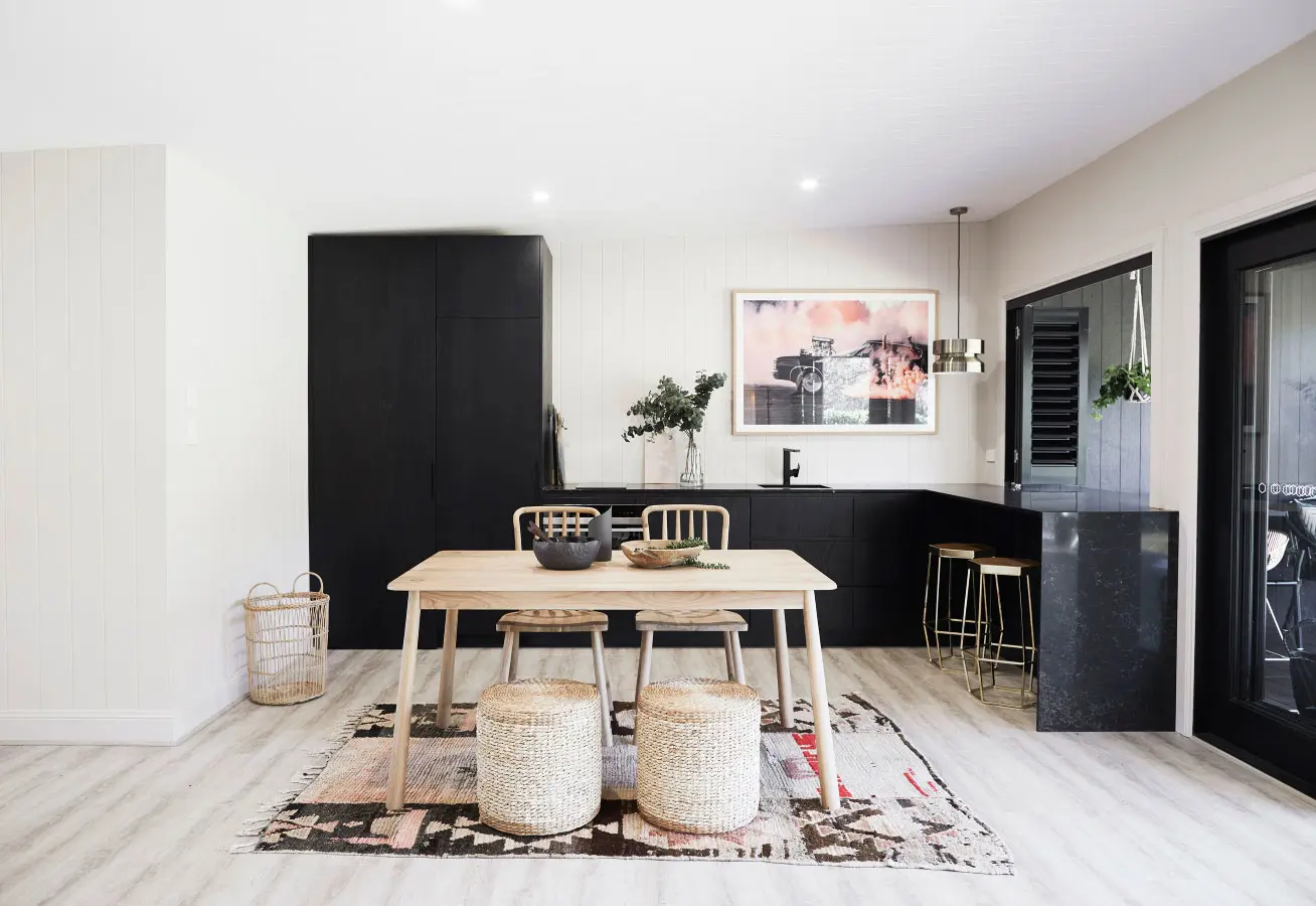 Kitchen with black cabinetry and a scandi style table and chairs. There are two stools at the table which is sitting on a patterned mat on a wooden floor.