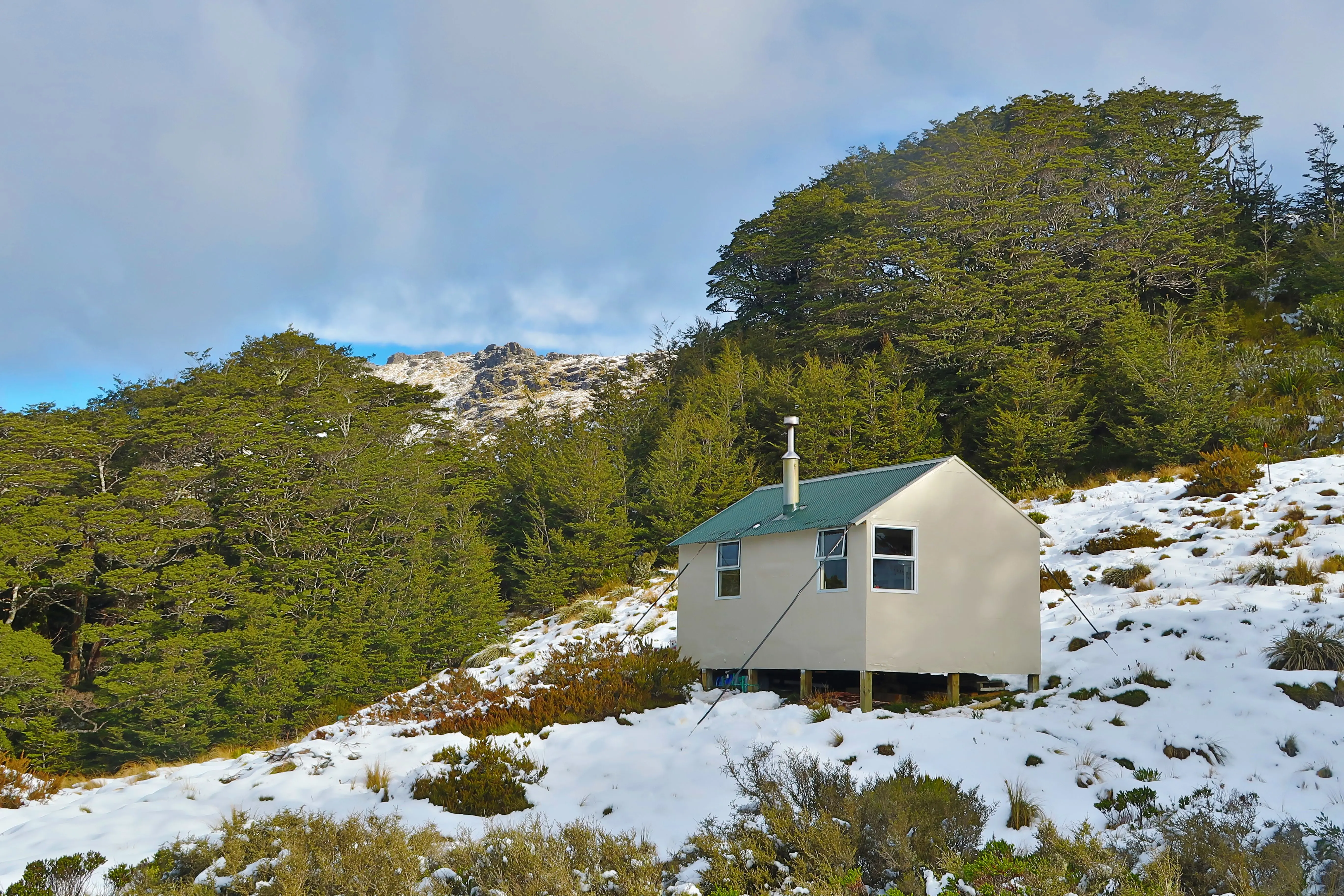 Mt Fell hut has a green roof and white exterior. It sits on a mountain covered in snow