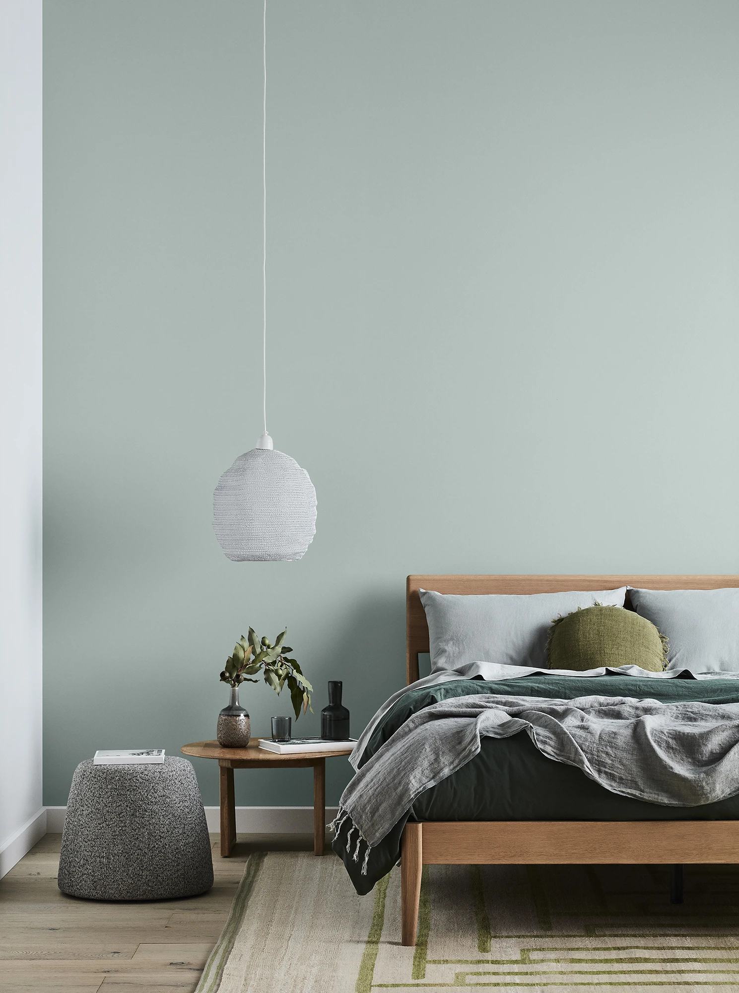 Bed in front of green wall. Side table with plant on it and a hanging white light