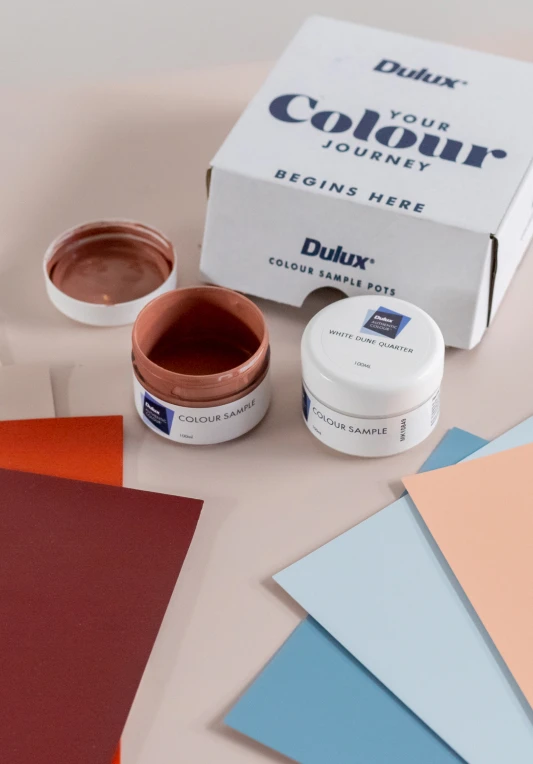 Two sample pots - one with it's lid off next to the box they came in which is Dulux branded and some colour swatches on a table
