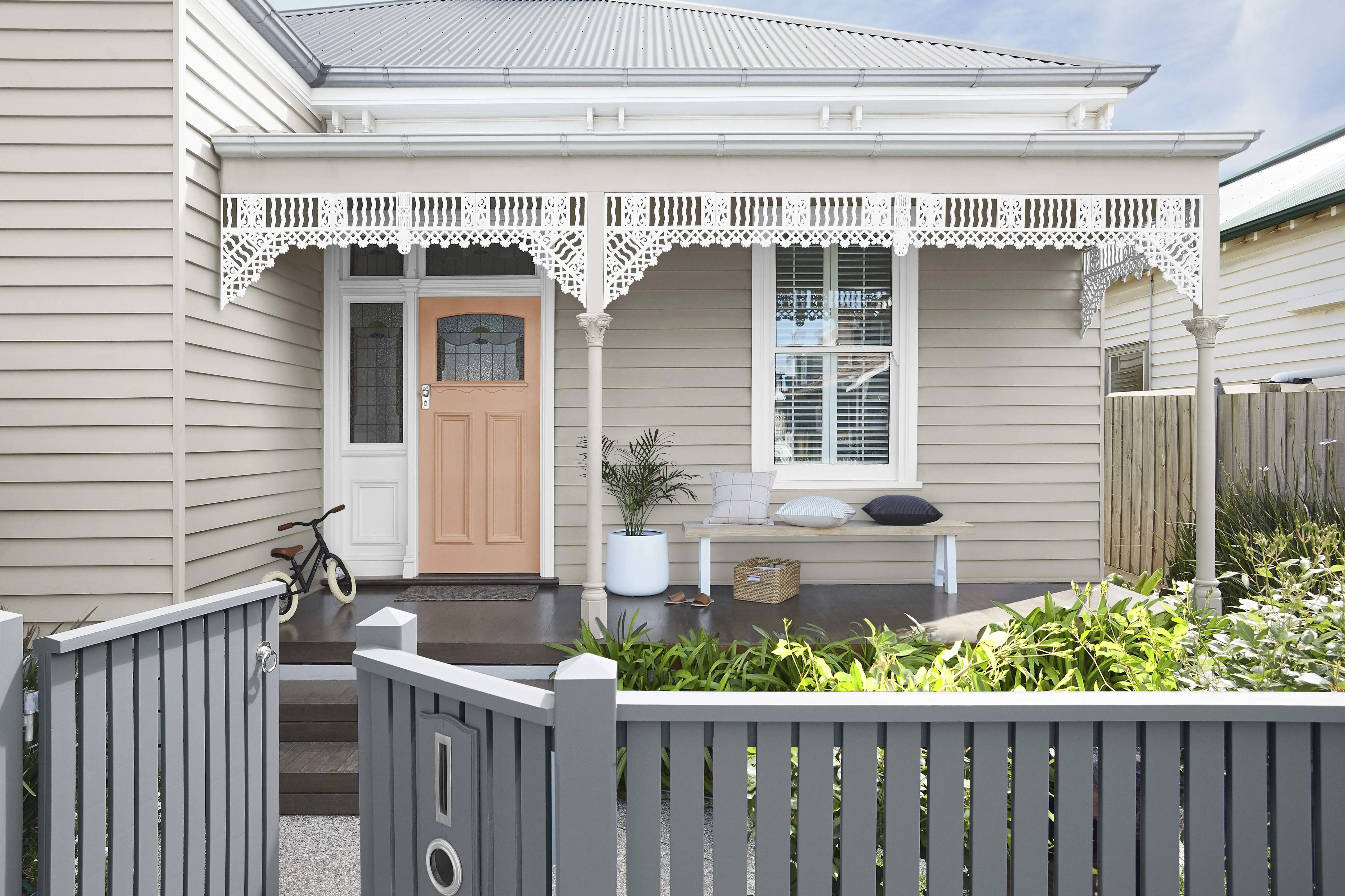 Neutral-coloured double-fronted Victorian weatherboard with white iron lace, grey picket fence and apricot door