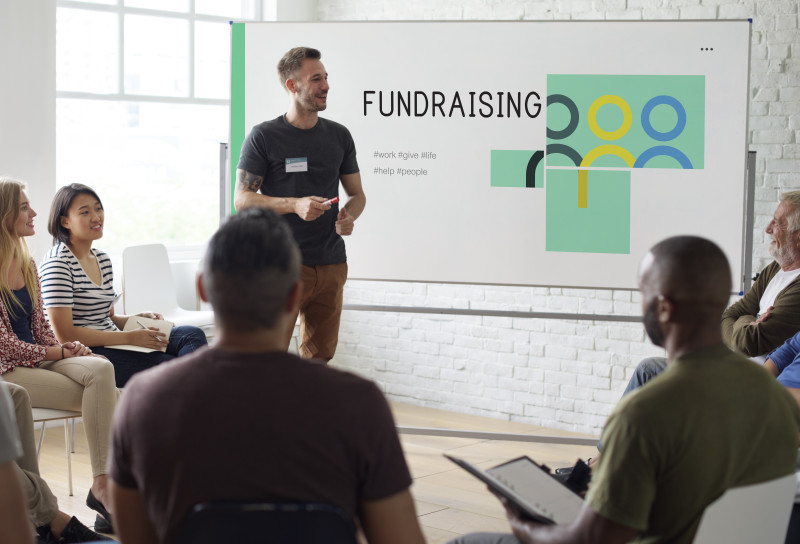A person standing and presenting in front of a group of people sitting down. Behind the person presenting is a white board that has 'FUNDRAISING' written on it.