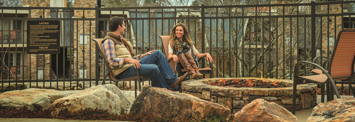 A young couple, dressed for autumn weather, sit beside the stone fire pit at The Residences at Biltmore.