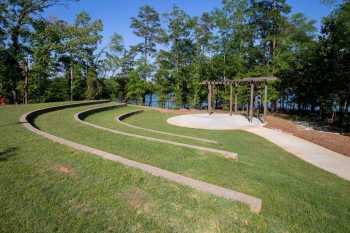 Three grassy tiers of amphitheater seating face a round, trellis-covered concrete pad, while beyond a narrow stand of trees, Lake Hartwell reflects a clear, blue sky.