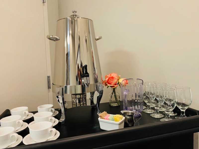 An assortment of glass stemware and espresso cups share a bar top with a stainless steel drink cooler and two pink roses.