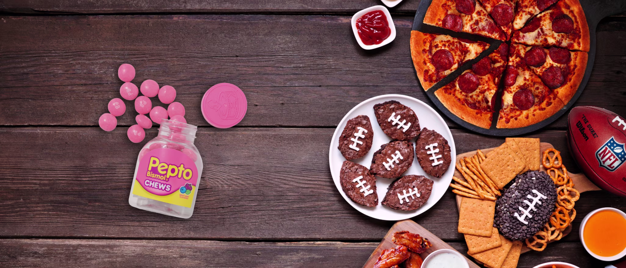 Open bottle of Pepto Bismol Chews spilling pink tablets on a wooden table beside football game snacks, including pepperoni pizza, chicken wings, pretzels, crackers, cookies, and football-shaped treats.