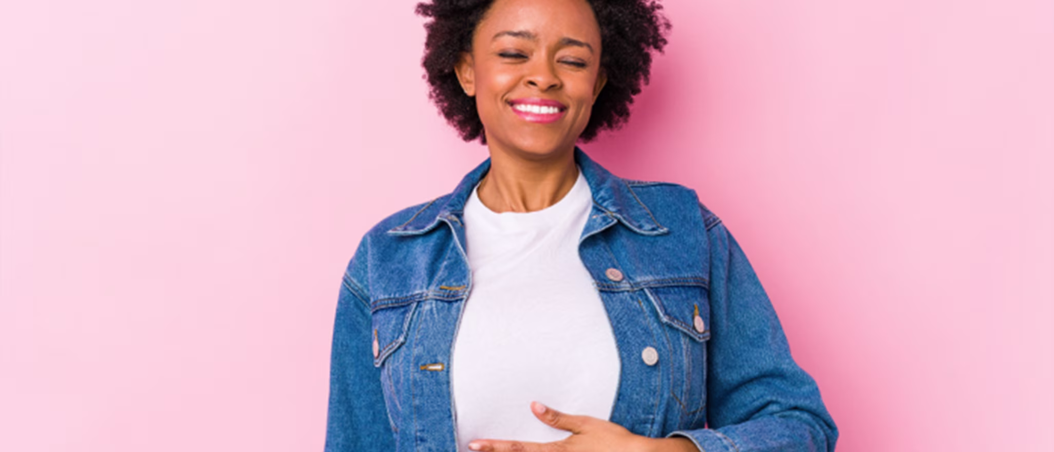 A woman in a denim jacket standing against a pink background, smiling and resting her hand on her stomach.