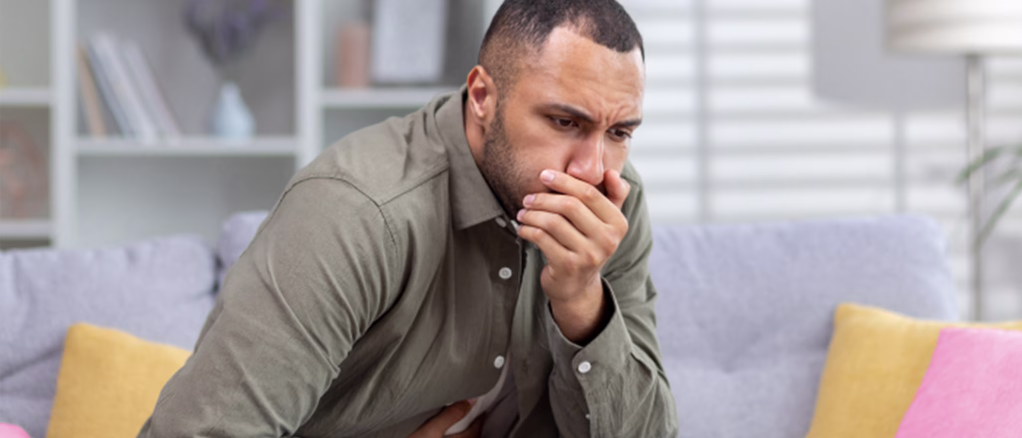 A man sitting on a sofa, looking nauseous with his hand over his mouth and the other on his stomach.