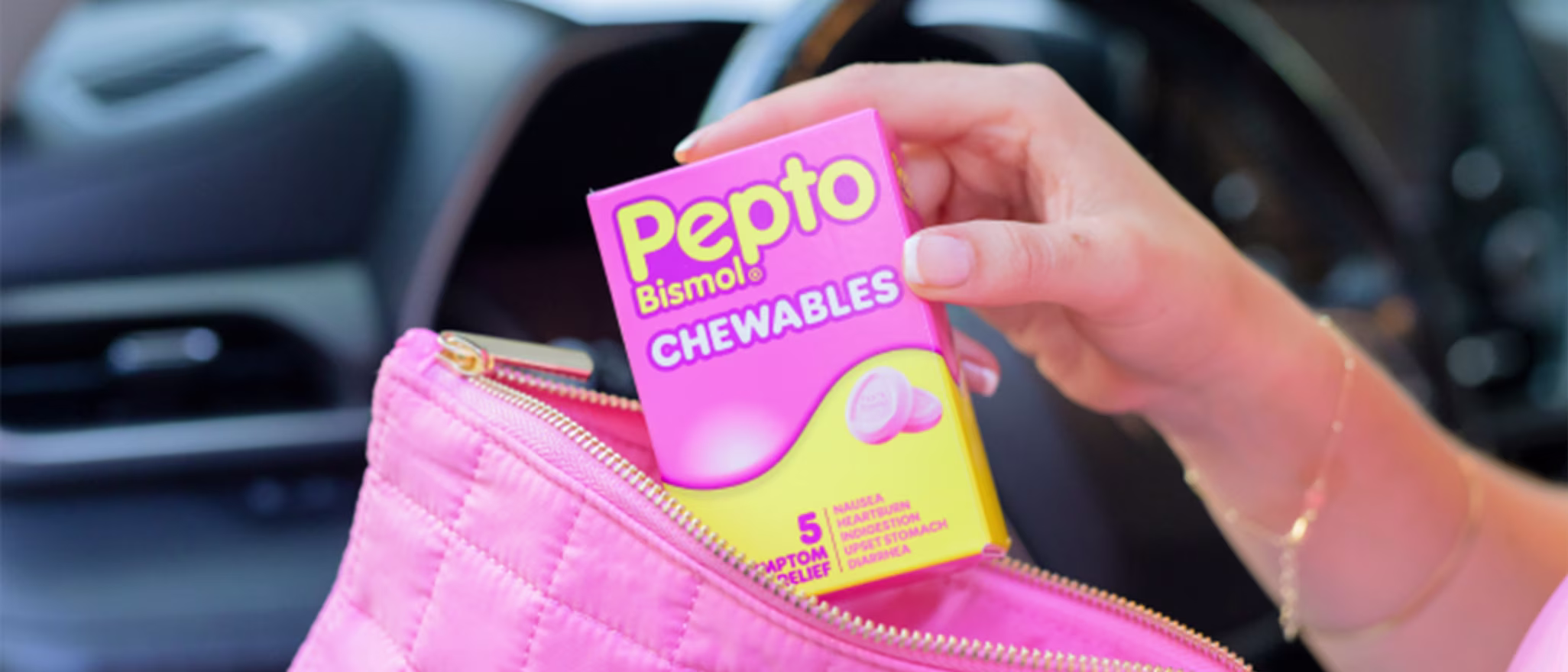 A hand placing a box of Pepto Bismol Chewables into a pink quilted makeup bag inside a car.